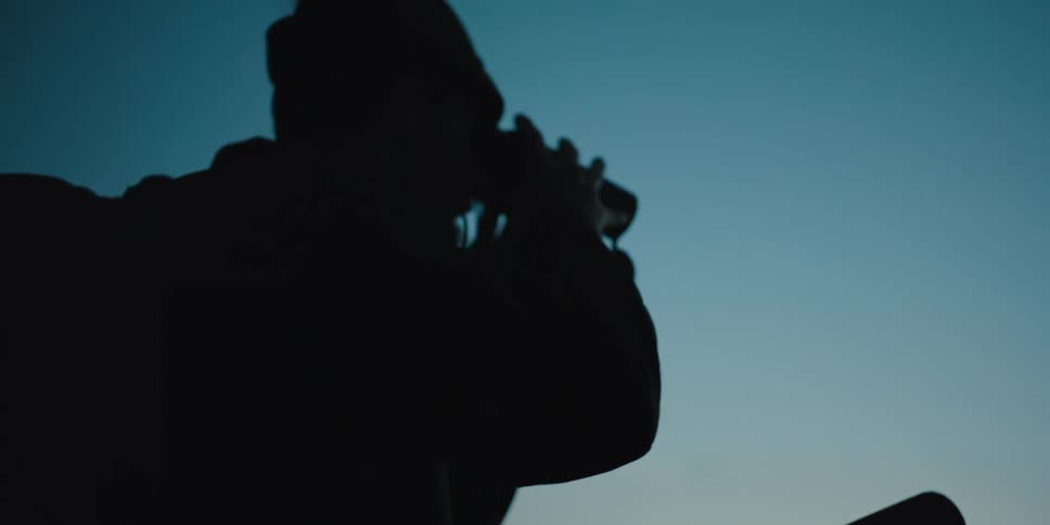 Hunter carrying a rifle looks through binoculars at sunset next to the mountains