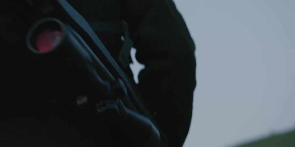 A person carrying a scoped rifle walks up a hill during the day
