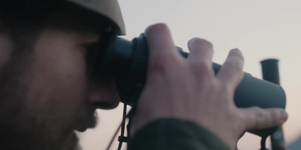 A man standing in the hills drives a staff into the ground before raising a pair of binoculars to his face