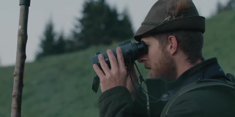 A man standing outside on a hill looks through binoculars