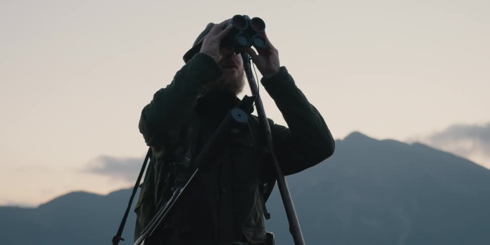 A man hiking with a dog while carrying a rifle over his shoulder raises a pair of binoculars to examine his surroundings before continuing forward
