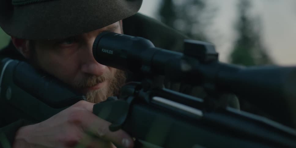 A man looks through the scope of a rifle while lying prone in the grass
