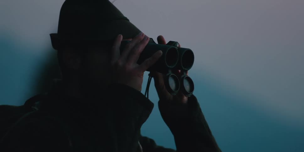 Man wearing hiking pack and hat lifts binoculars to eyes