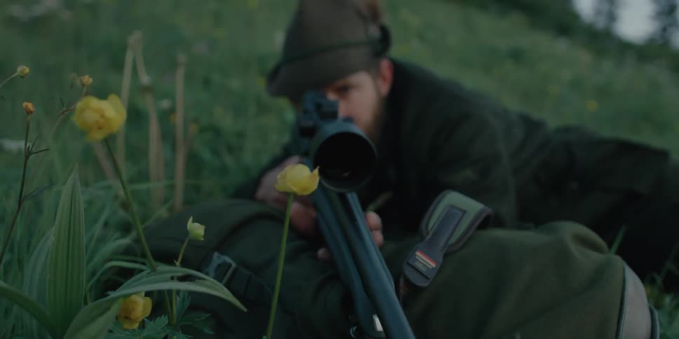 A man rests his rifle on his backpack as he takes aim while lying in a field of grass
