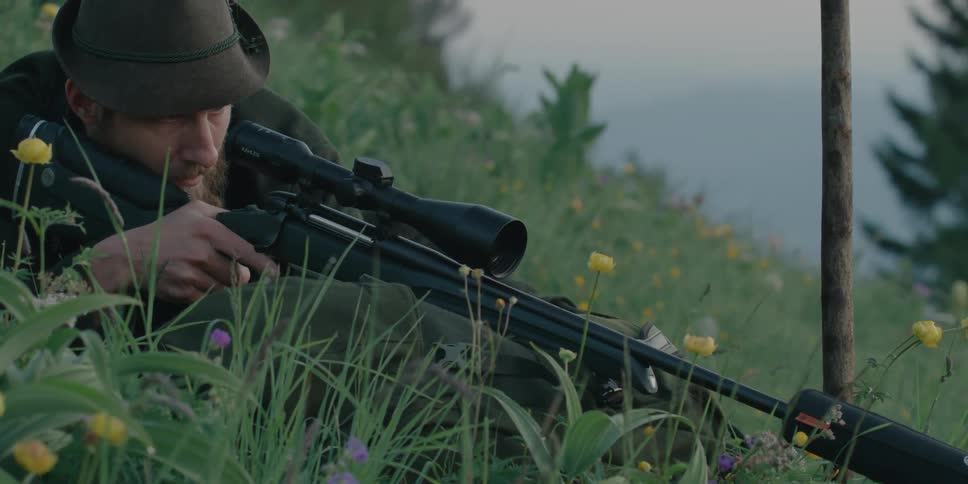 A young man lying on a hill points his gun and gets ready to shoot
