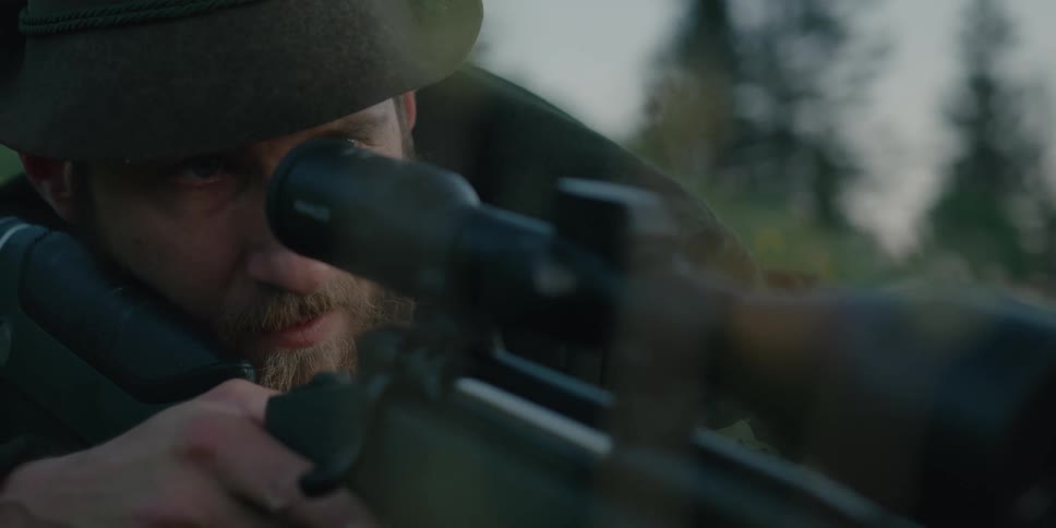 A man with a hat aiming a rifle with trees and the sky in the background