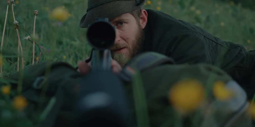 A man lies in the grass on the side of a hill while looking down the scope of a long rifle