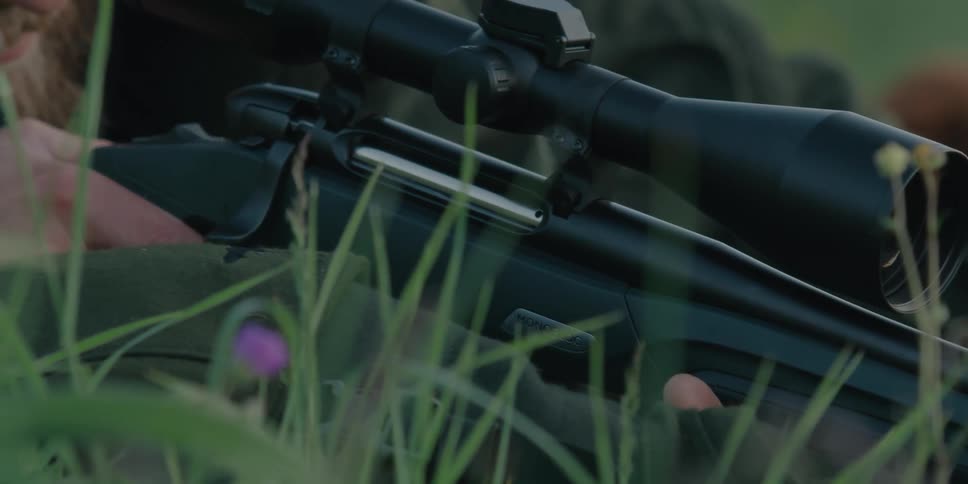 A dog lies in the grass next to a man looking through the scope of a rifle