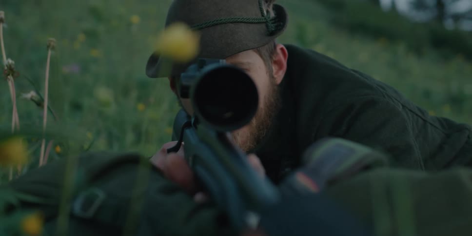 Hunter lying on the grass in a hill looks through the scope mounted on a rifle