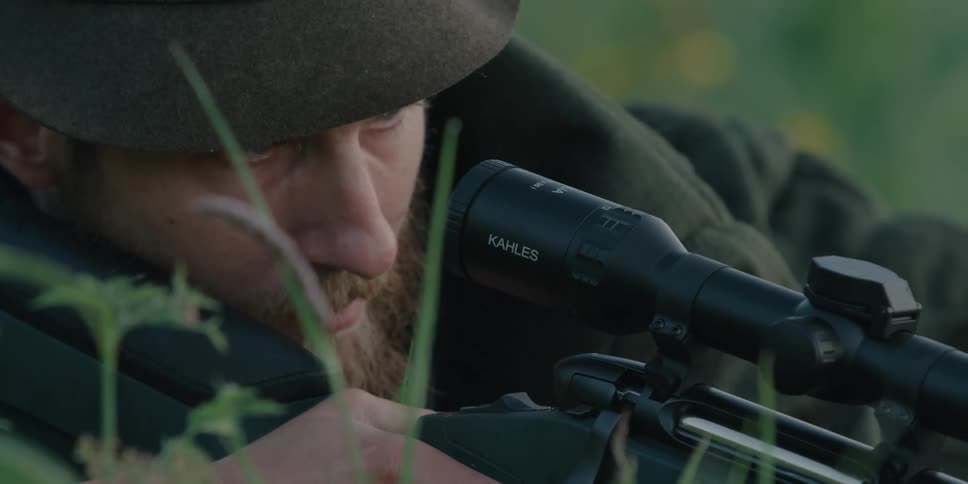 A white man with a beard aims a rifle with scope