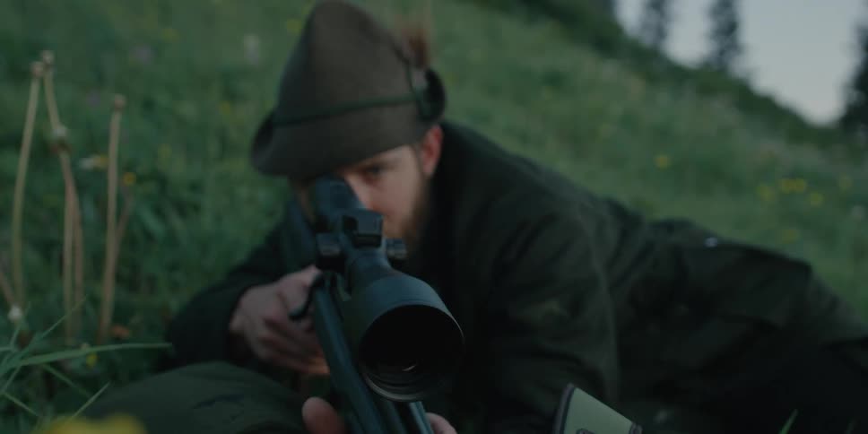 A man lies on the ground in a field and looks through the scope of his rifle