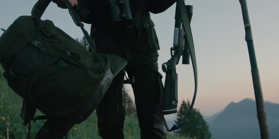 A man holding a gun puts his backpack down on a hill as he waits for his prey