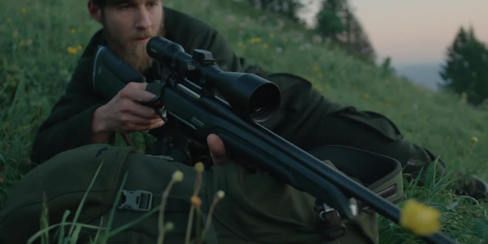 A hunter lays down on the grass placing a rifle over a backpack at morning