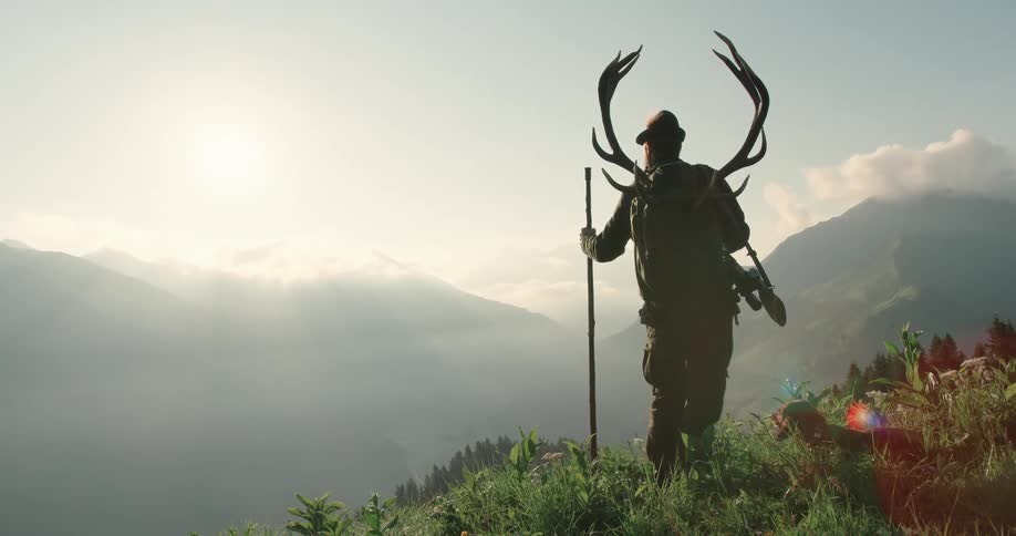 Man carrying elk antlers and gun stands above mountain range with dog