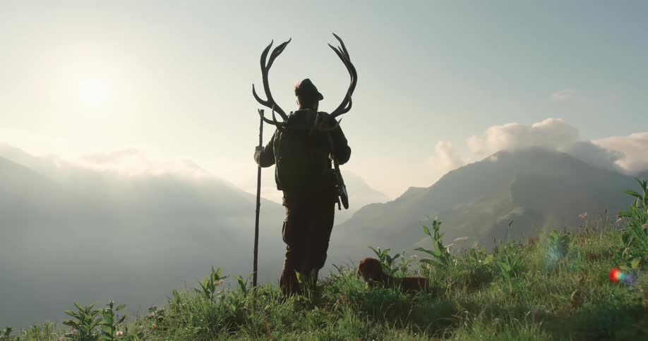 Man stands looking at foggy mountains next to dog while carrying antlers on back