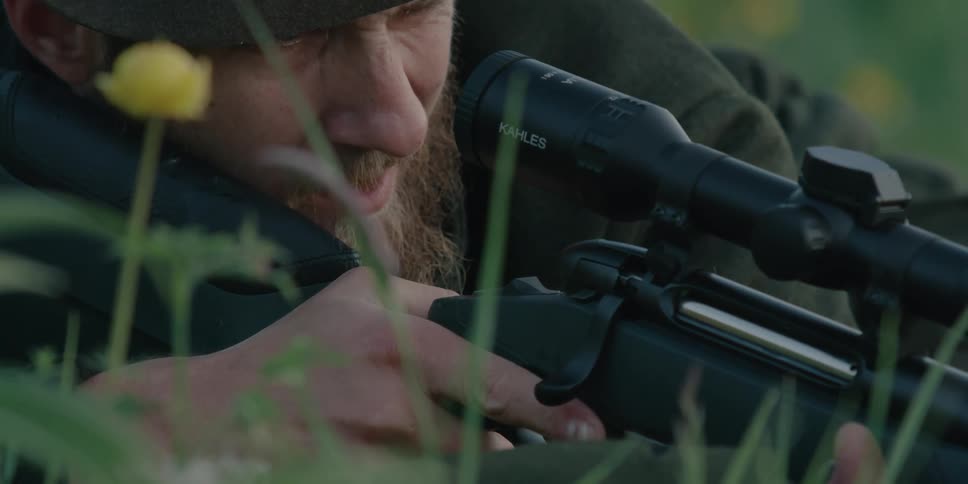 A bearded man lying prone in the grass checks his rifle before looking down the sights
