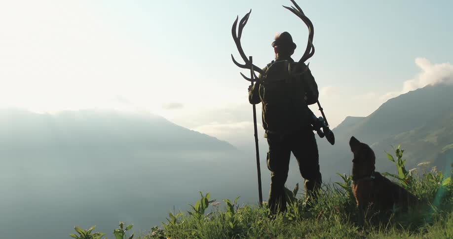 A man with antlers strapped on his back stand on a hill with his dog looking for his prey