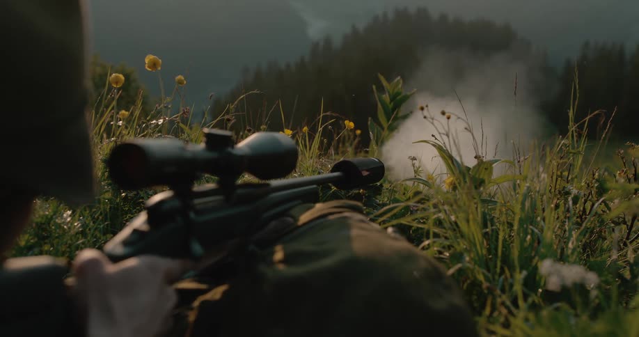 Hunter shoots a rifle while laying on the grass on a hill in the mountains