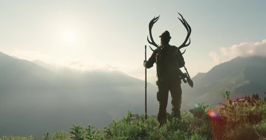 Hunter with deer antlers in a backpack stands atop a hill near the mountains with his dog