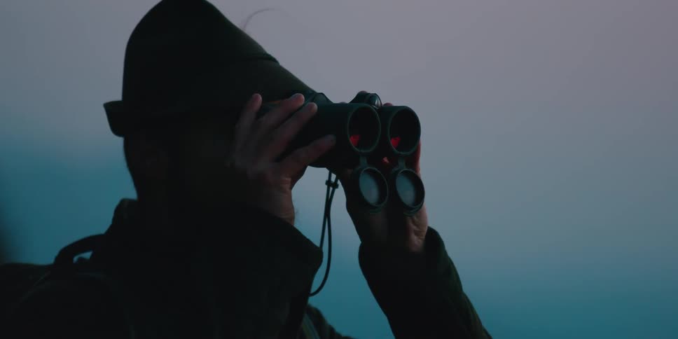 A man standing outside with a gun lifts up his binoculars and looks through them and then walks away
