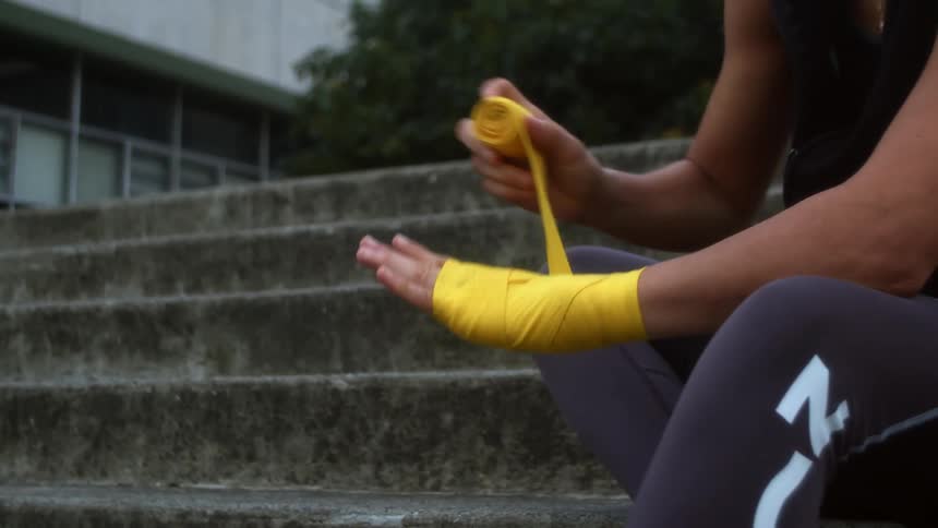 A woman sits on a cement staircase outdoors as she wraps her hands with yellow hand wrap