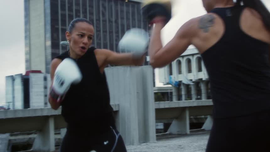 Two women train using boxing gloves and focus pads on a city rooftop