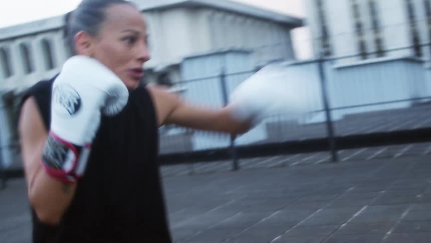 Two women on a rooftop training with boxing gloves with buildings and the sky in the background