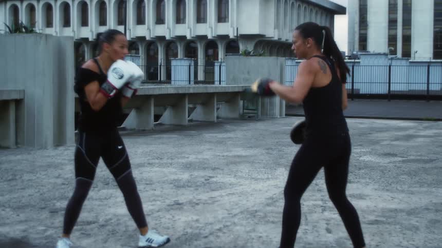 Two women on a rooftop training with boxing gloves with buildings in the background