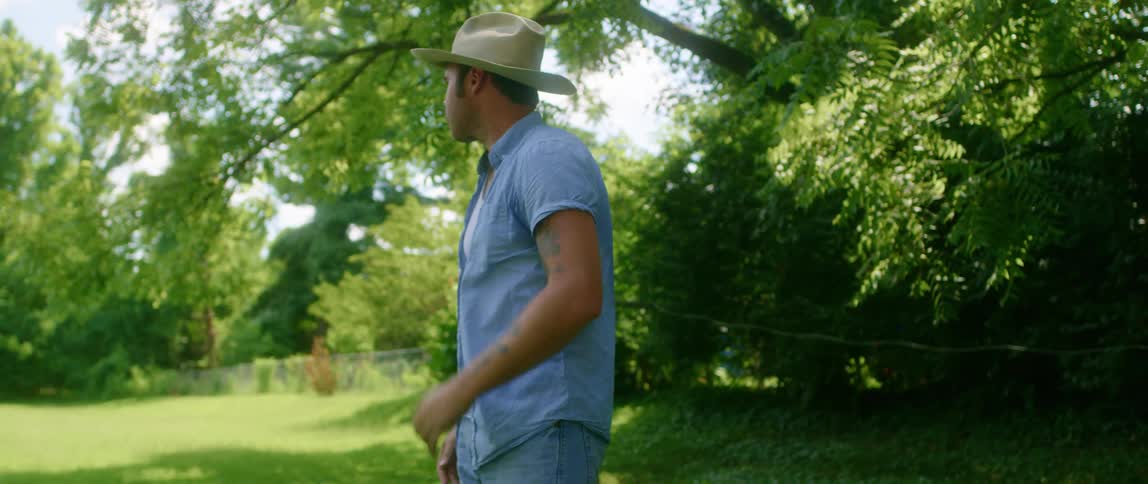 Man standing outside wearing denim shirt and jeans smooths hair and puts on cowboy hat