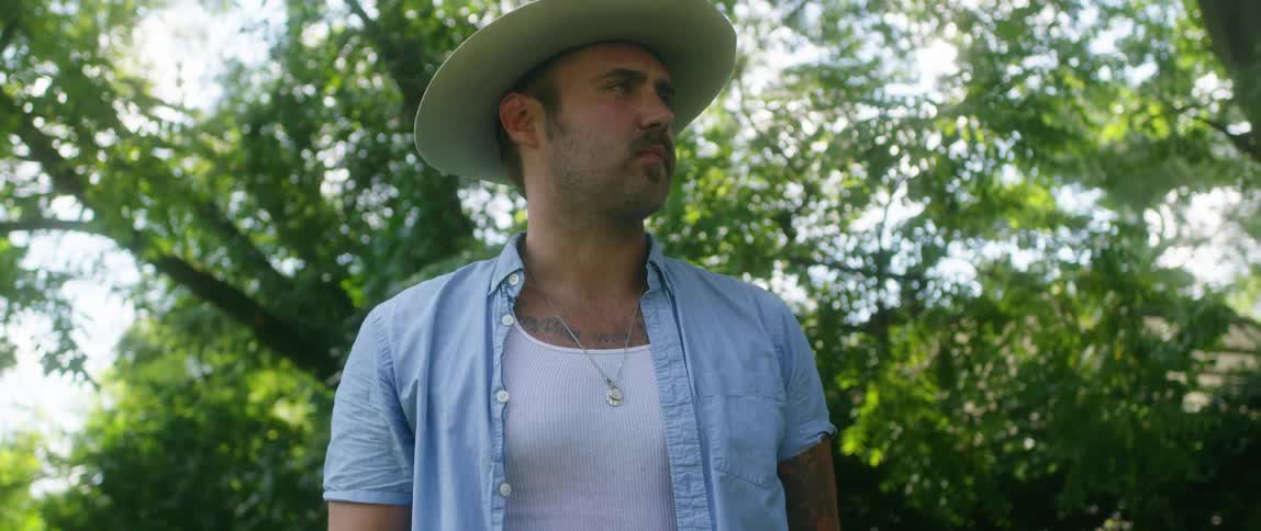 A man in a cowboy hat waits in front of a tree before walking away