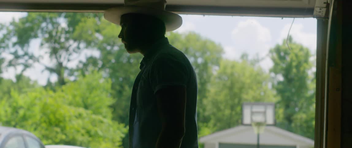 A man stands in his garage and places a cowboy hat on his head