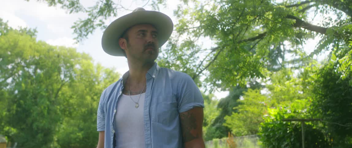A man with a hat standing on a grassy field with trees and the sky in the background