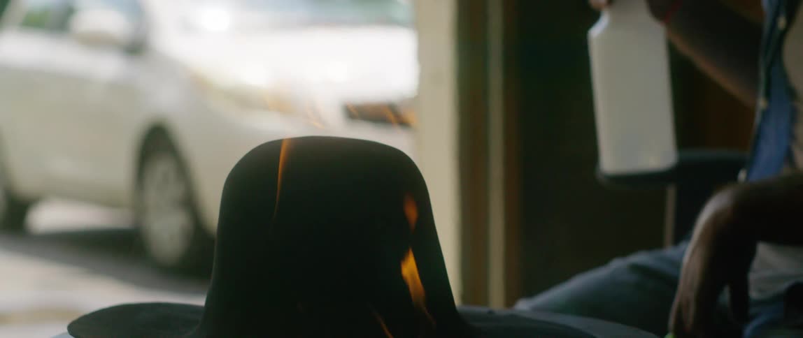 A man sits in his garage in front of a hat that is on fire and sprays water on it