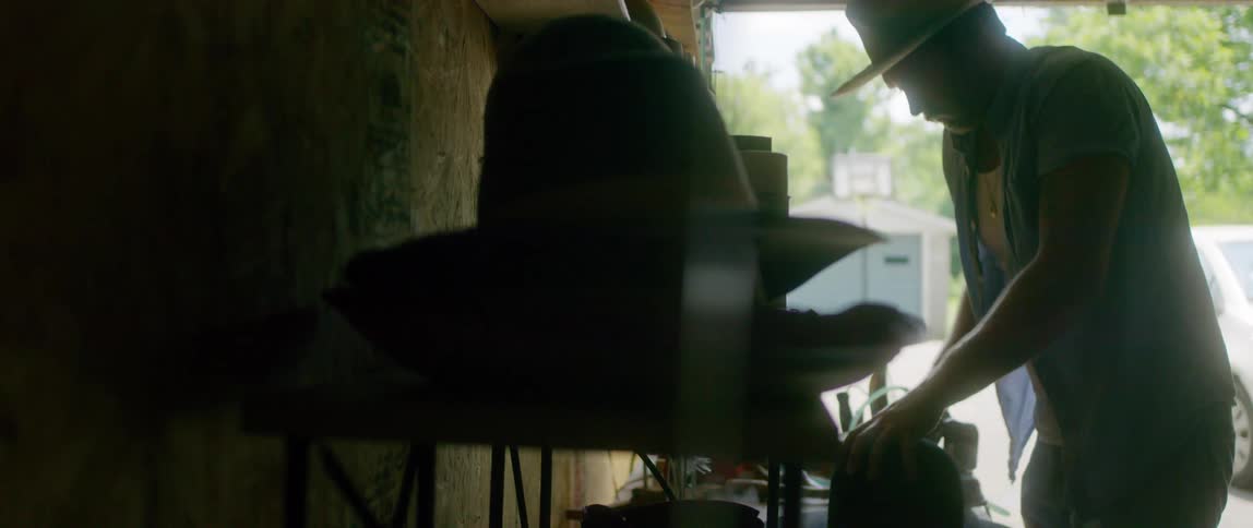 A man in his garage flattens the brim of his hat with an iron