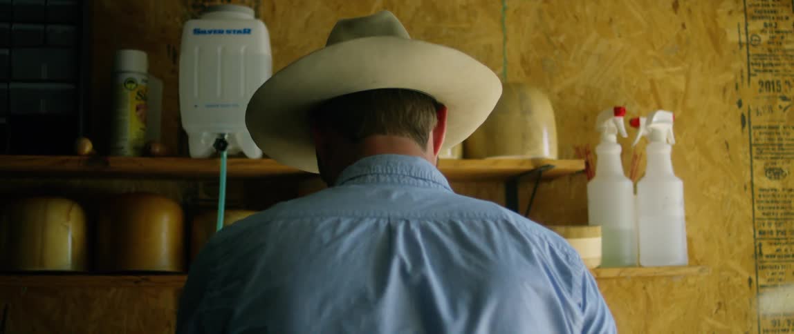 A man holds a frying pan as he stands in front of two shelves in a room with particle board walls