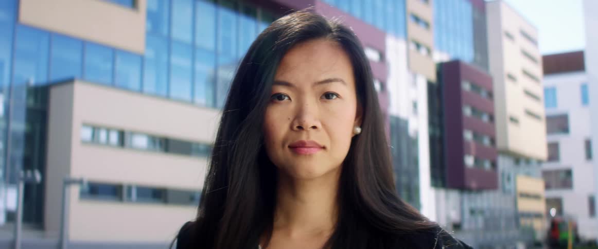 A middle aged asian woman stares into the camera while standing outdoors in a metropolis