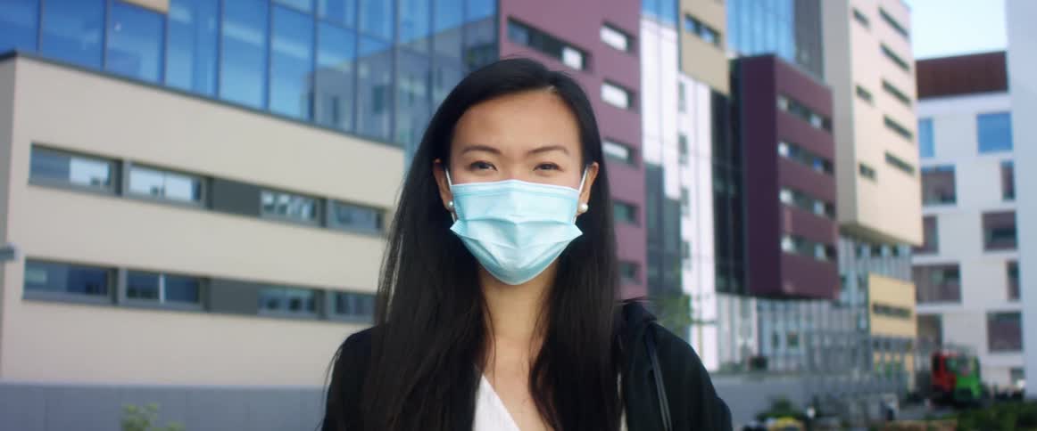 A woman wearing a medical mask stands outside large buildings alone