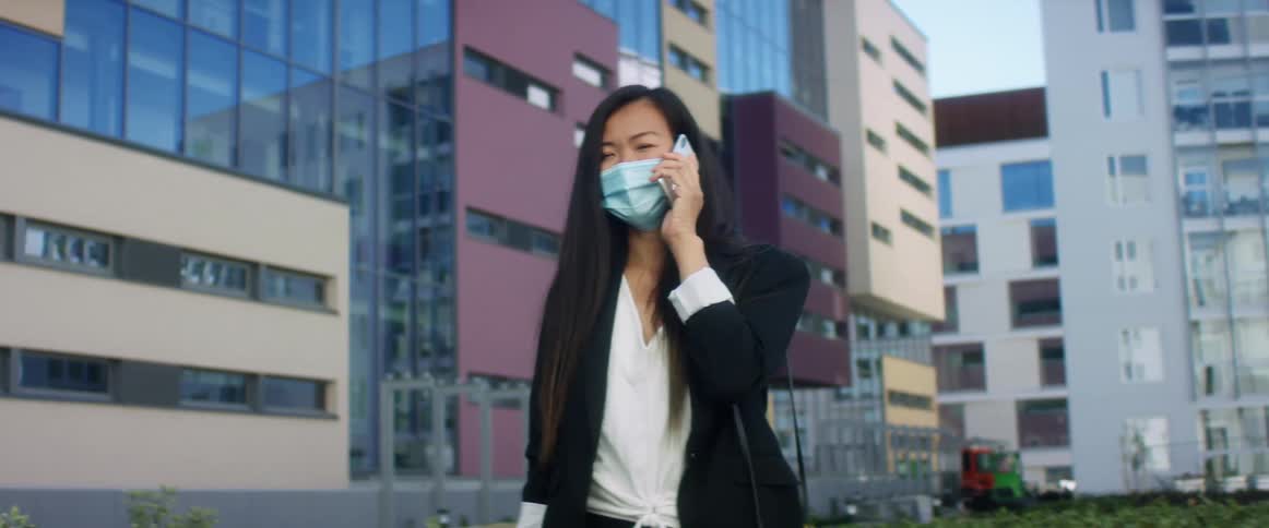 Asian woman with with long hair and a blue mask talks on the smartphone in a city street with tall buildings in the background