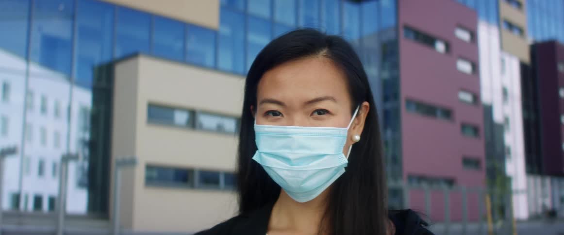 Asian woman with long straight hair and a blue maskfaces the camera with eye contact