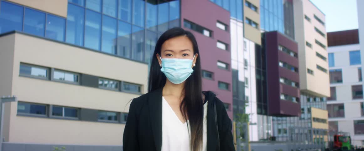 A woman standing outside of office buildings in a downtown area puts a mask on