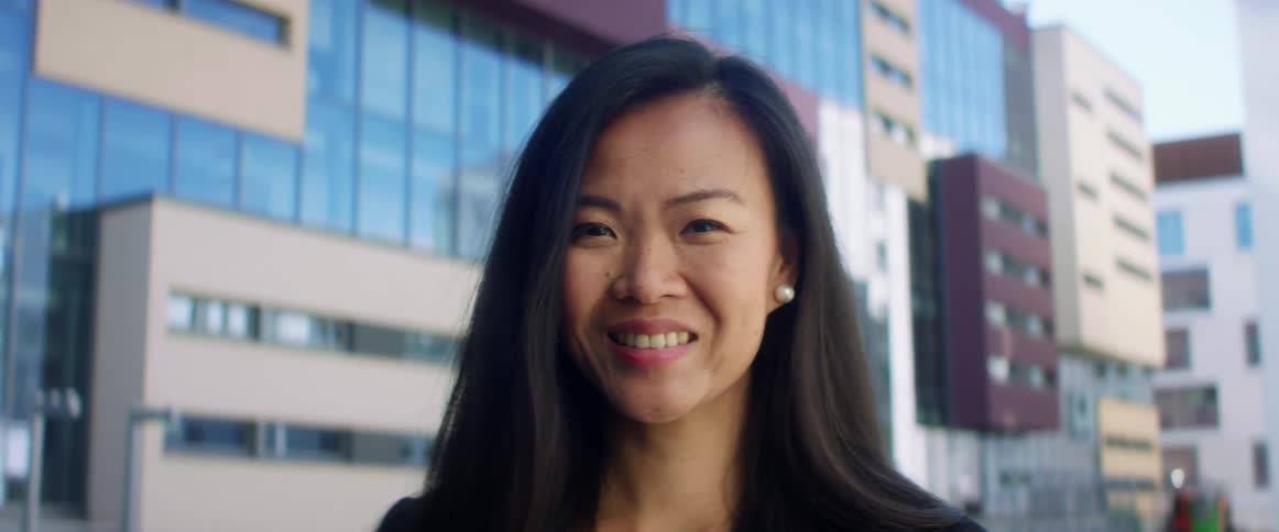 An asian woman smiles into the camera while standing outdoors in a metropolis during the day