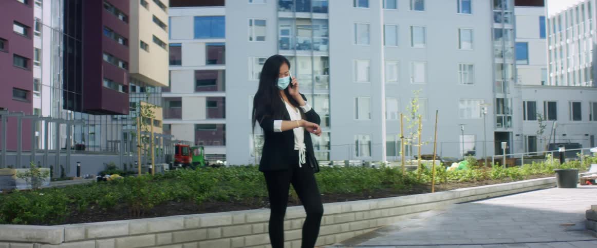 A woman stands outside a large city building talking on a cell phone and looking at her watch