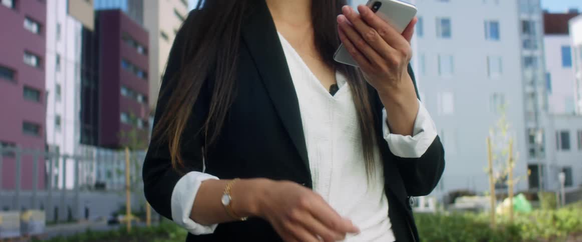 An asian woman takes a smartphone from her purse and raises it to begin typing while standing outdoors in a metropolis during the day