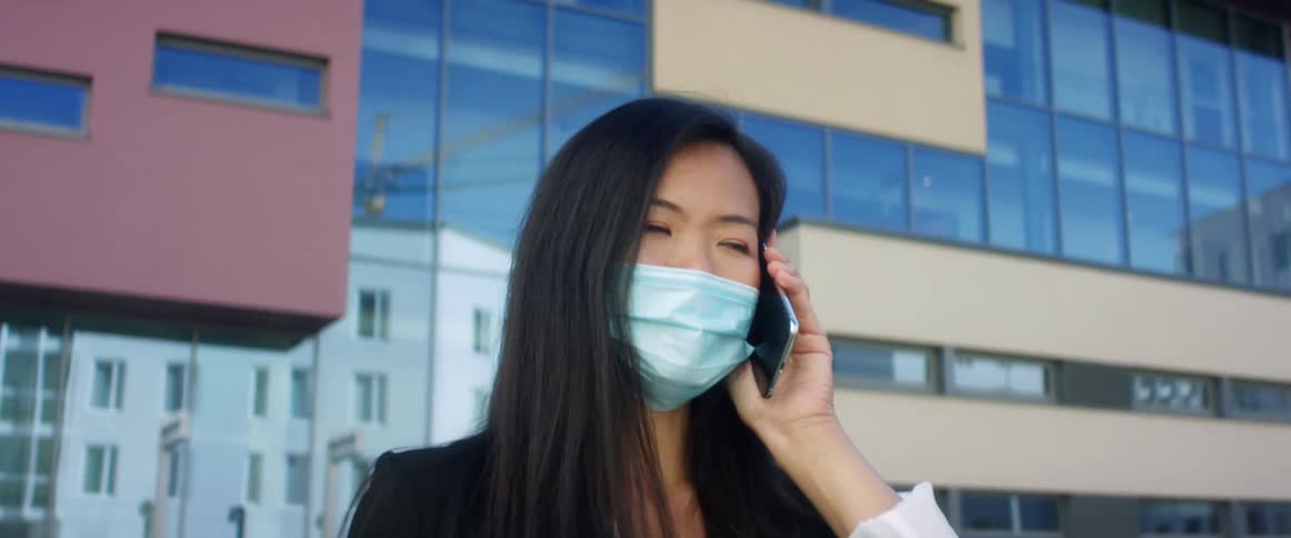 Woman wearing face mask talks on mobile phone near building reflecting other buildings in its windows