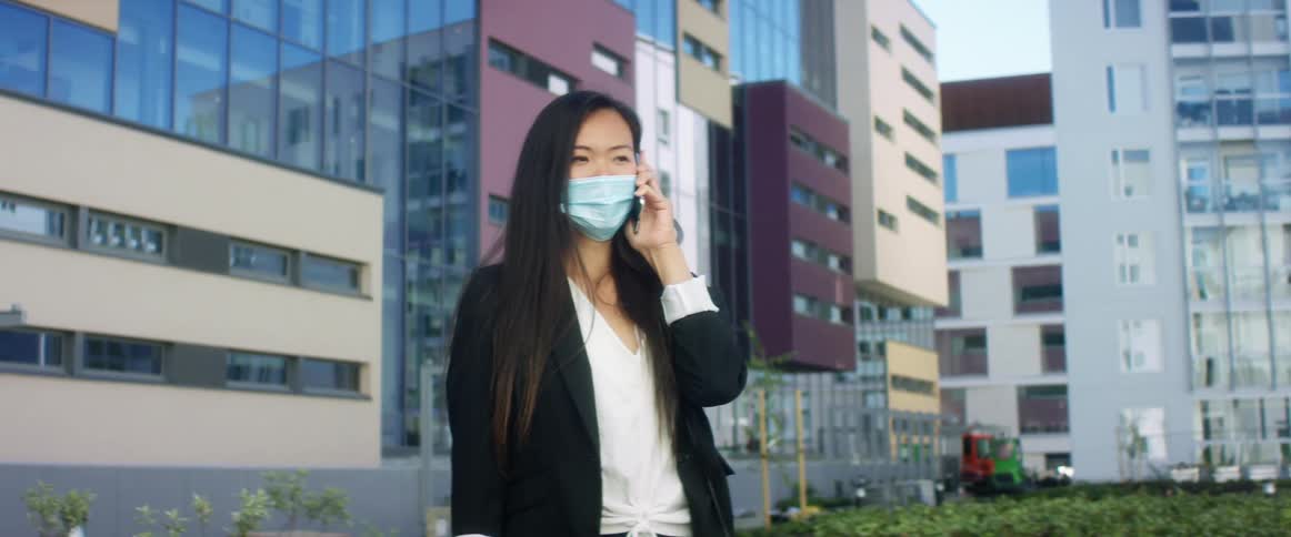 Woman in business casual clothing and face mask makes phone call outside modern office building