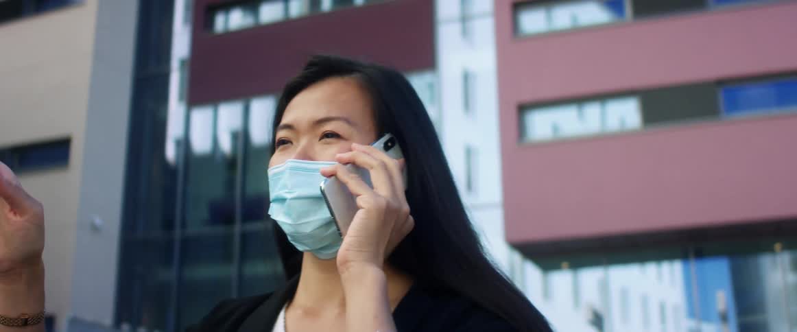 Asian woman with long hair and blue mask talks on smartphone in front of high rise buildings