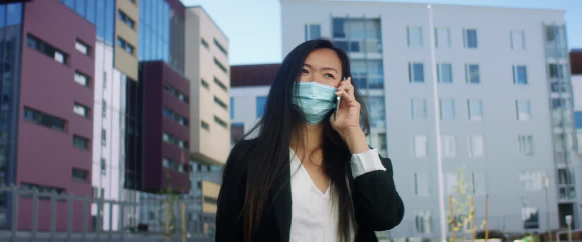 Asian woman with long hair and a blue mask talks on the smartphone while looking at her wristwatch in the street with apartments in the background