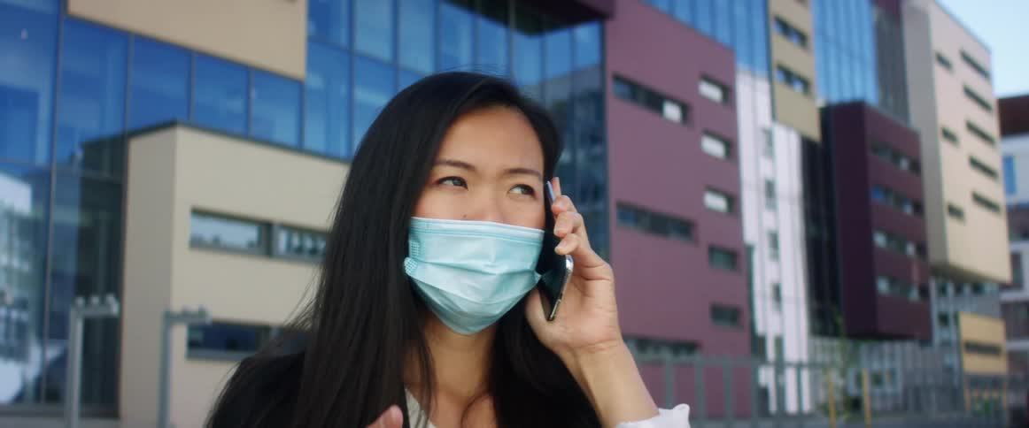 Woman wearing a face mask talks on her smart phone outside a building in the city