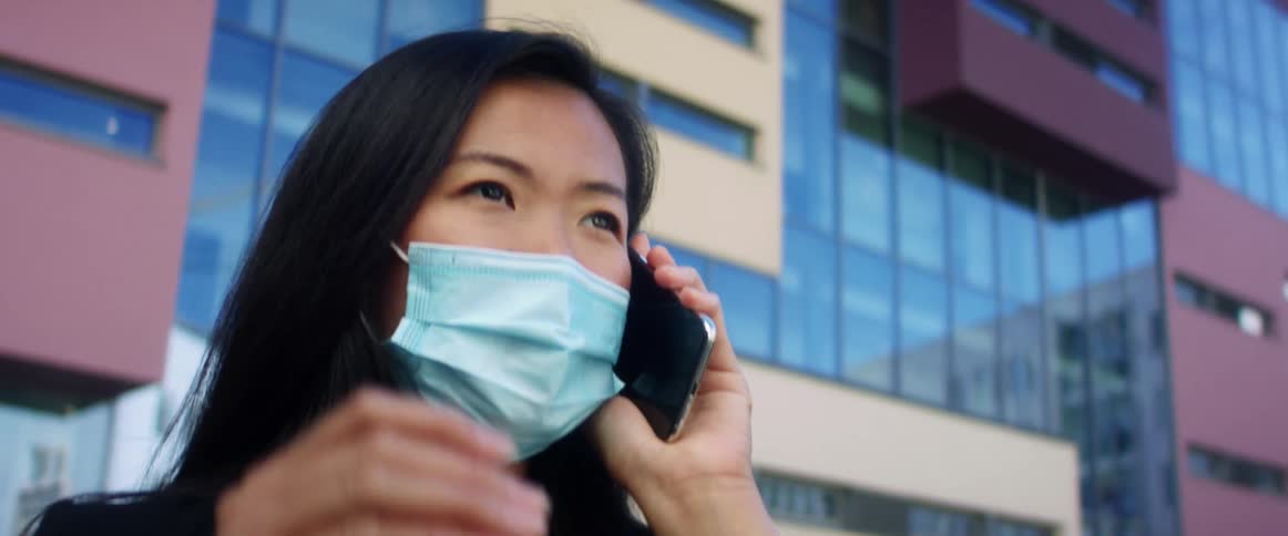 Asian woman with long hair and a blue mask talks on the smartphone in the street