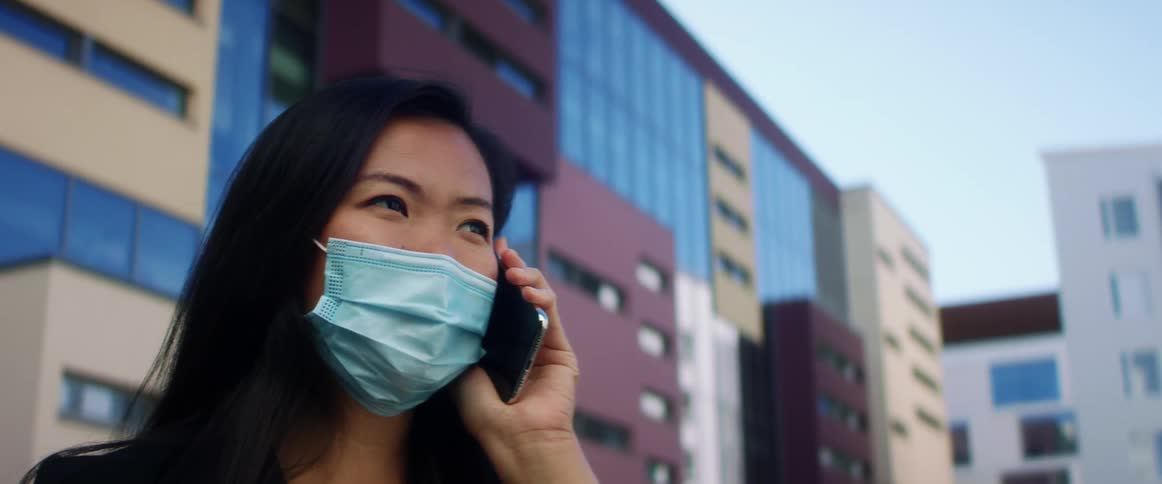 Asian woman with with long hair and a blue mask talks on the smartphone in a city street